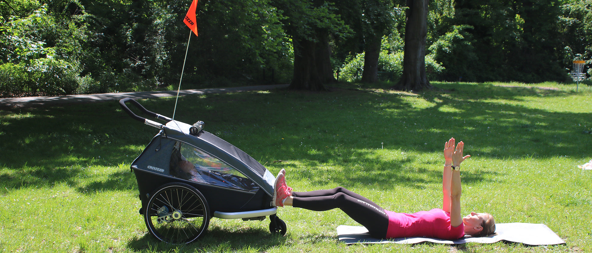 Woman performing bridge exercise with feet on Croozer bike trailer and arms raised on yoga mat in park